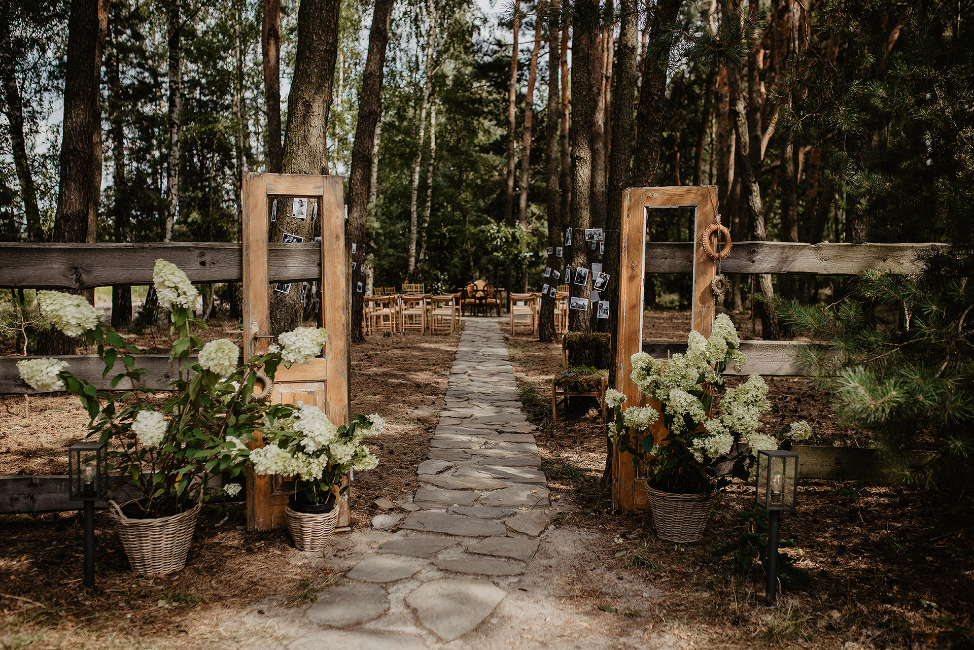 Malownicza ścieżka prowadząca do miejsca ceremonii ślubnej w lesie Sielsko Anielsko, ozdobiona starymi drzwiami, kwiatami i zdjęciami, sfotografowana przez Camera Folks – Wedding Photo & Video.