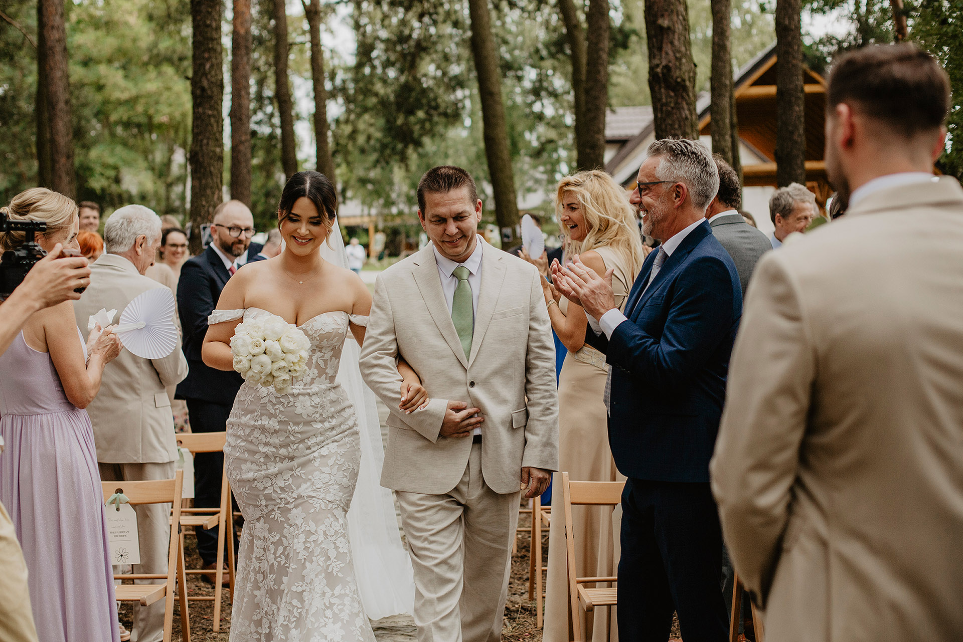 Panna Młoda w koronkowej sukni ślubnej i z bukietem, idąca z uśmiechem wzdłuż rzędu gości w lesie Sielsko Anielsko podczas ceremonii zaślubin, sfotografowana przez Camera Folks – Wedding Photo & Video.