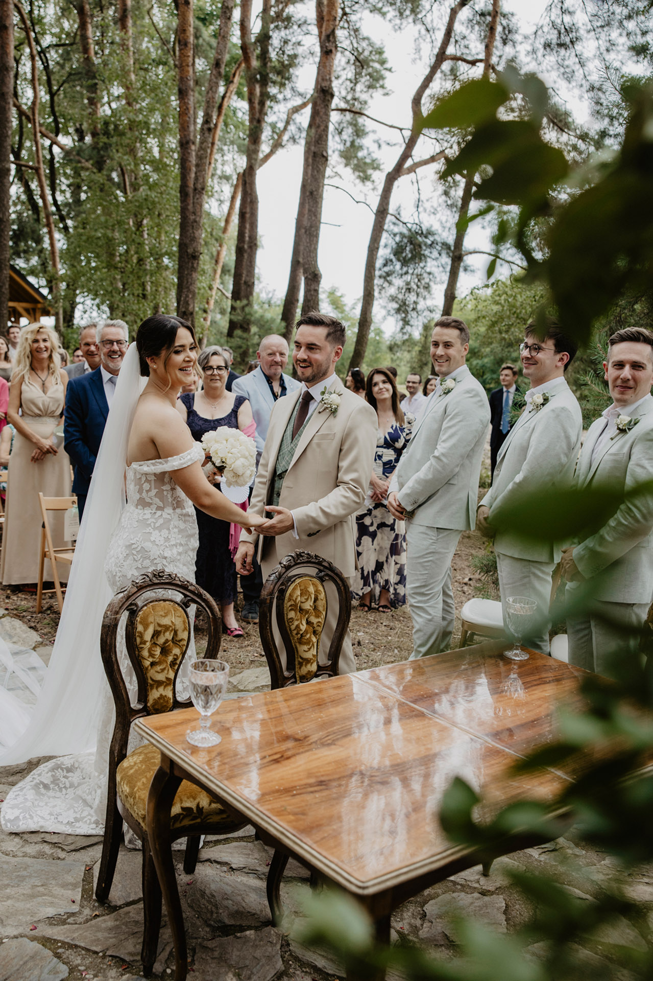 Panna Młoda i Pan Młody, uśmiechnięci, trzymający się za ręce przy stole ceremonii, otoczeni gośćmi w leśnym otoczeniu Sielsko Anielsko, sfotografowani przez Camera Folks – Wedding Photo & Video.