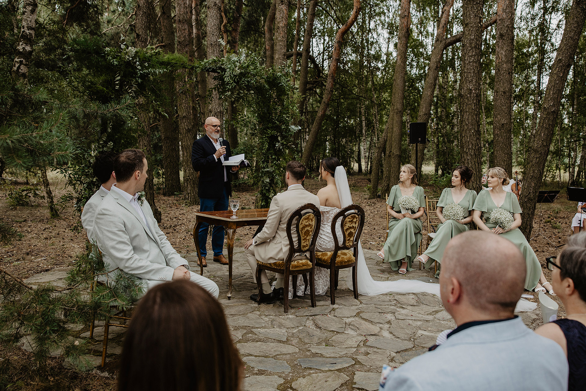 Para Młoda siedząca przy stole ceremonii w lesie Sielsko Anielsko, z pastorem prowadzącym uroczystość i gośćmi, sfotografowana przez Camera Folks – Wedding Photo & Video.