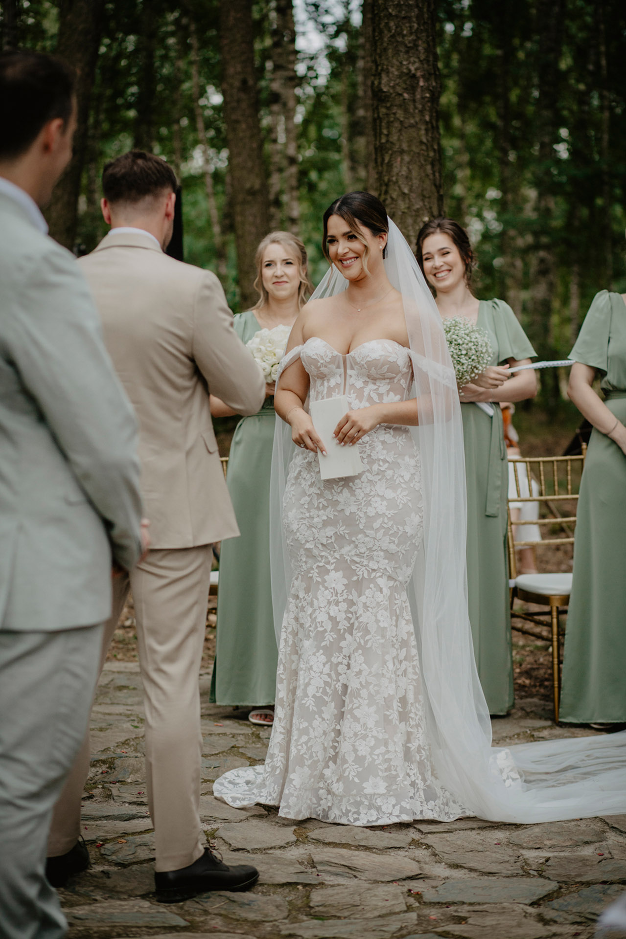 Panna Młoda w koronkowej sukni ślubnej, z uśmiechem i kartką w dłoni, stojąca naprzeciwko Pana Młodego podczas ceremonii w plenerze Sielsko Anielsko, sfotografowana przez Camera Folks – Wedding Photo & Video.