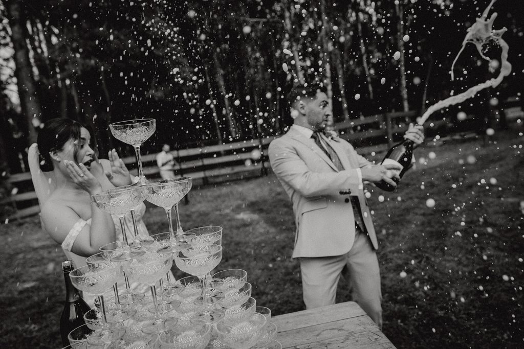 Para Młoda podczas "champagne tower" – Pan Młody otwiera szampana, który rozbryzguje się wokół, a Panna Młoda reaguje z zaskoczeniem i radością, w Sielsko Anielsko, czarno-białe zdjęcie by Camera Folks – Wedding Photo & Video.