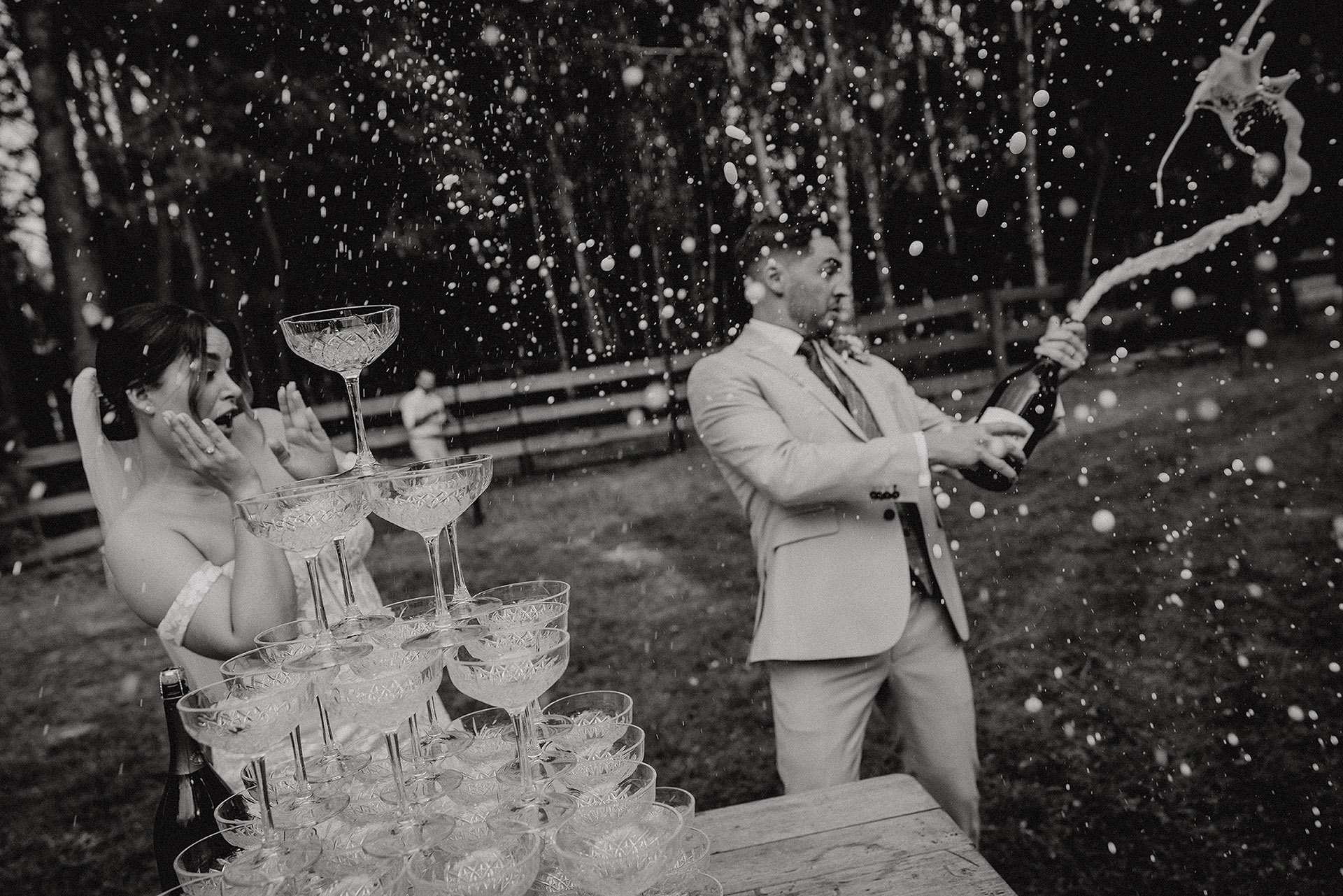 Para Młoda podczas "champagne tower" – Pan Młody otwiera szampana, który rozbryzguje się wokół, a Panna Młoda reaguje z zaskoczeniem i radością, w Sielsko Anielsko, czarno-białe zdjęcie by Camera Folks – Wedding Photo & Video.
