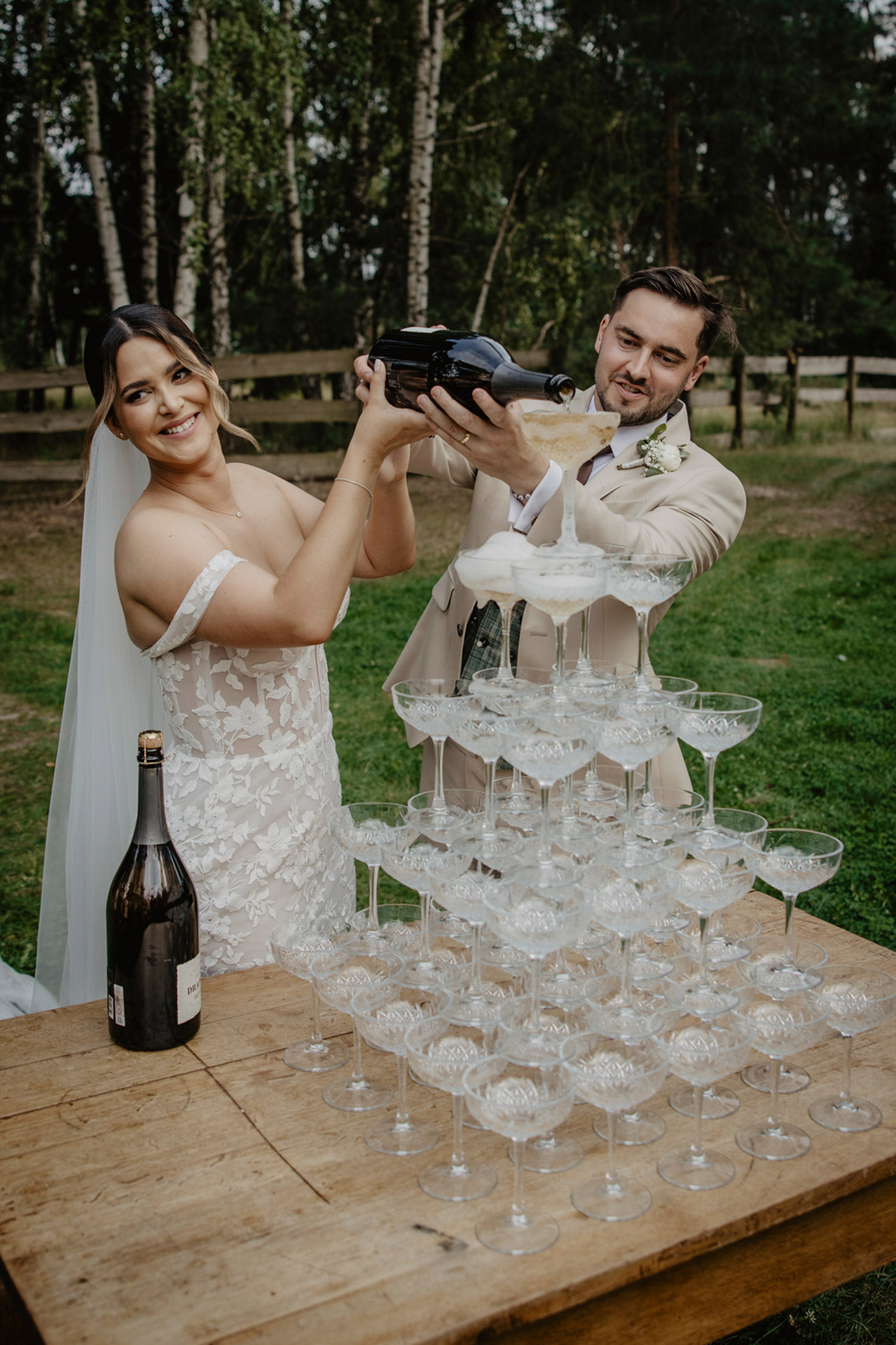 Para Młoda uśmiechnięta, wylewająca szampana do wieży kieliszków podczas "champagne tower" w Sielsko Anielsko, sfotografowana przez Camera Folks – Wedding Photo & Video.