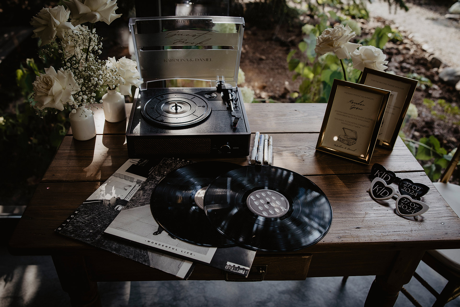 Oryginalna księga gości weselnych w formie gramofonu i płyt winylowych na drewnianym stole, z białymi kwiatami i okularami imprezowymi, w Sielsko Anielsko, sfotografowana przez Camera Folks – Wedding Photo & Video.