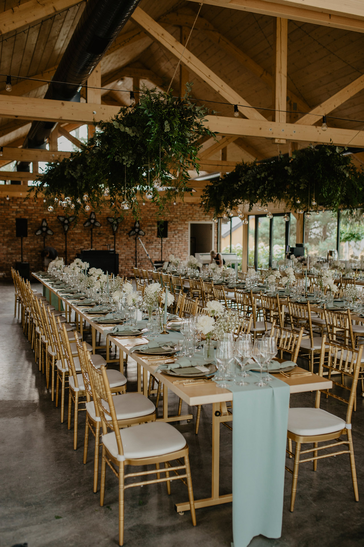 Elegancko udekorowane wnętrze nowoczesnej szklanej stodoły w Sielsko Anielsko, z długimi stołami i wiszącymi zielonymi dekoracjami, przygotowane na wesele przez Pracownię Rosochate, sfotografowane przez Camera Folks – Wedding Photo & Video.