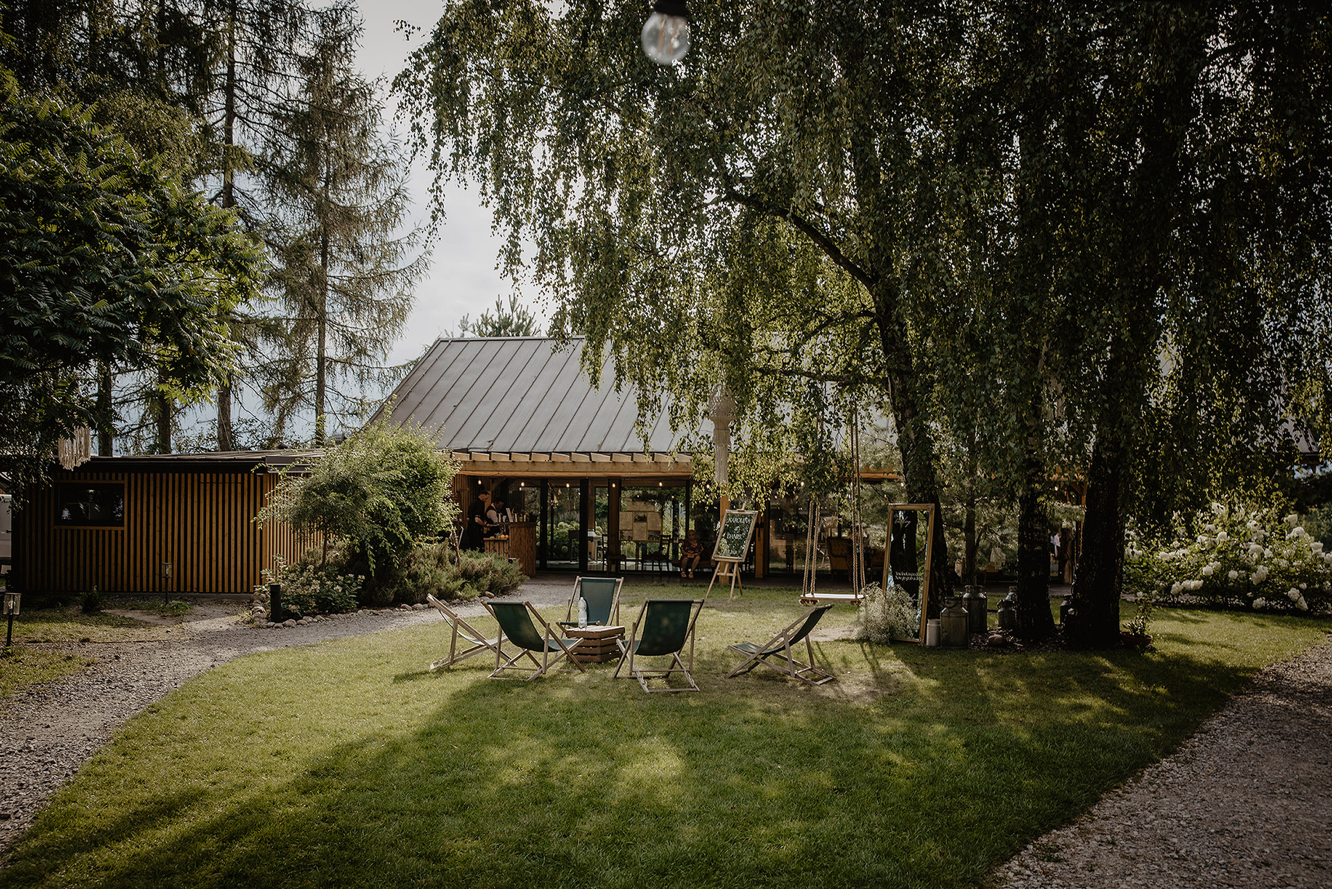 Widok na nowoczesną drewnianą stodołę weselną Sielsko Anielsko z metalowym dachem, otoczoną drzewami i zielonym trawnikiem z leżakami, sfotografowany przez Camera Folks – Wedding Photo & Video.