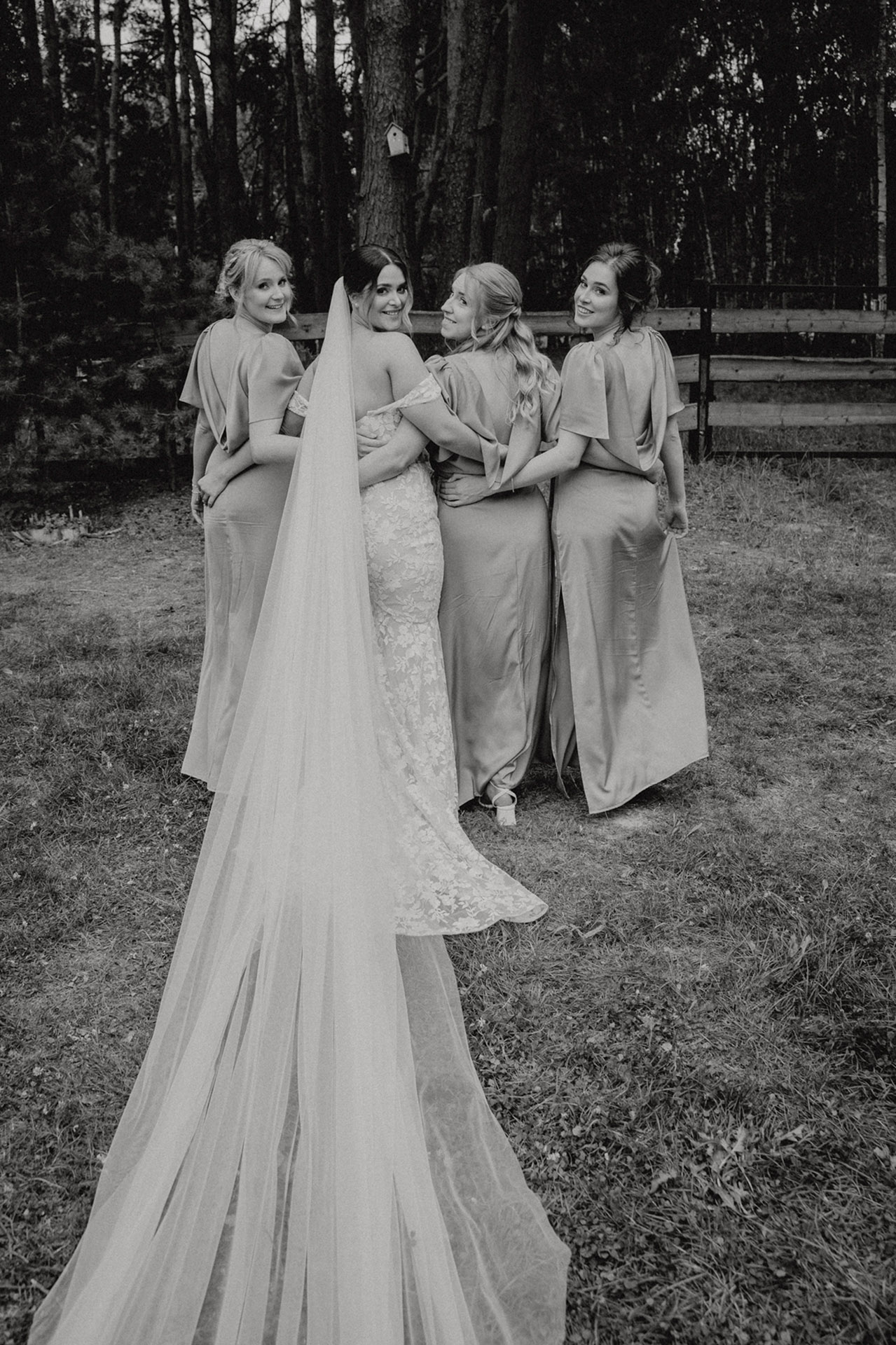 Czarno-białe zdjęcie Panny Młodej w długim welonie stojącej tyłem, otoczonej czterema druhnami w jasnych sukniach, na trawiastym terenie w Sielsko Anielsko, sfotografowane przez Camera Folks – Wedding Photo & Video.