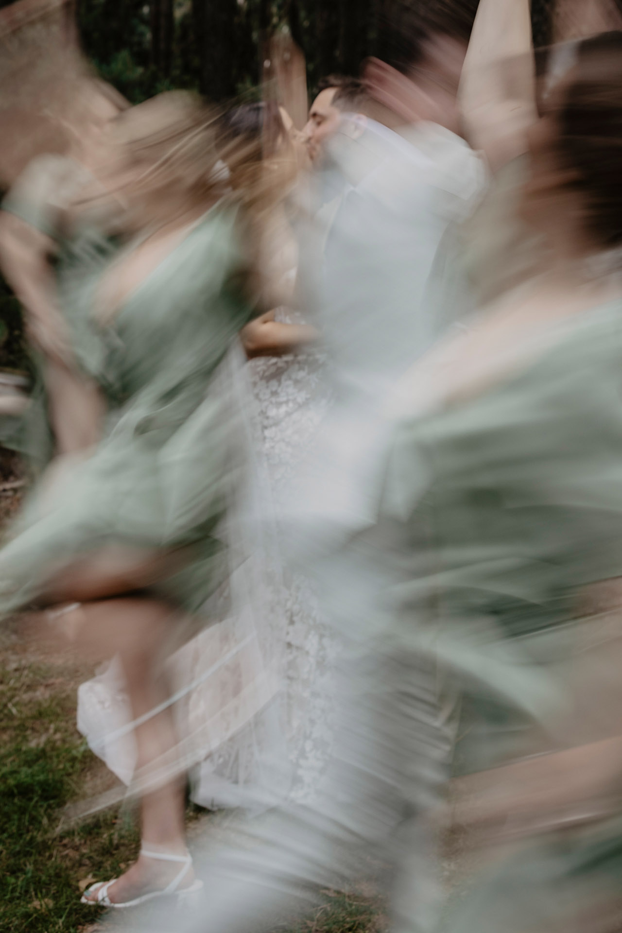 Artystyczne, rozmyte czarno-białe zdjęcie z efektem ruchu, przedstawiające Parę Młodą otoczoną tańczącymi druhnami i drużbami w jasnych ubraniach, w leśnym otoczeniu, sfotografowane przez Camera Folks – Wedding Photo & Video.