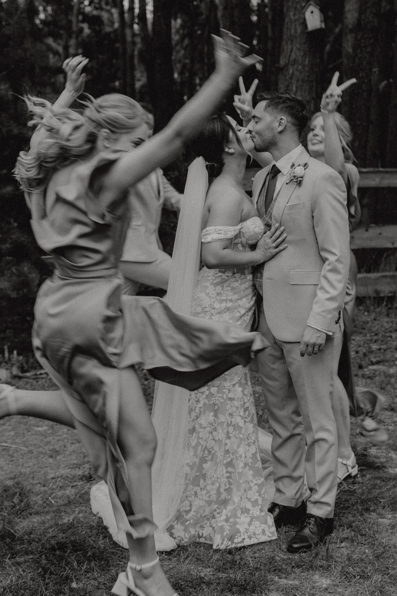 Czarno-białe, dynamiczne zdjęcie grupowe Panny Młodej i Pana Młodego, z uśmiechniętymi druhnami i drużbami, z których jedna osoba skacze i podnosi ręce, tworząc zabawny efekt w tle leśnym Sielsko Anielsko, sfotografowane przez Camera Folks – Wedding Photo & Video.