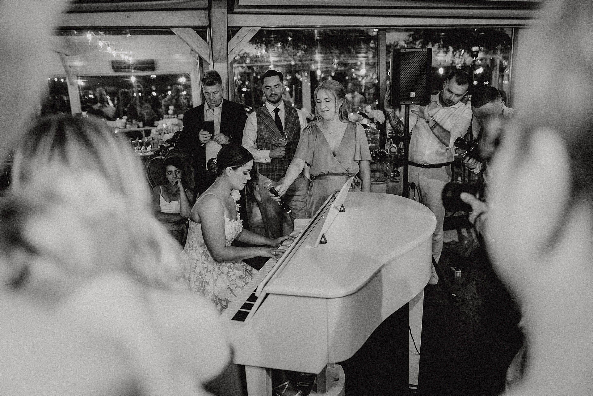 Czarno-białe zdjęcie Panny Młodej w sukni ślubnej śpiewającej i grającej na białym fortepianie, ujęcie zza rozmytych sylwetek gości, podczas przyjęcia weselnego w Sielsko Anielsko, sfotografowane przez Camera Folks – Wedding Photo & Video.