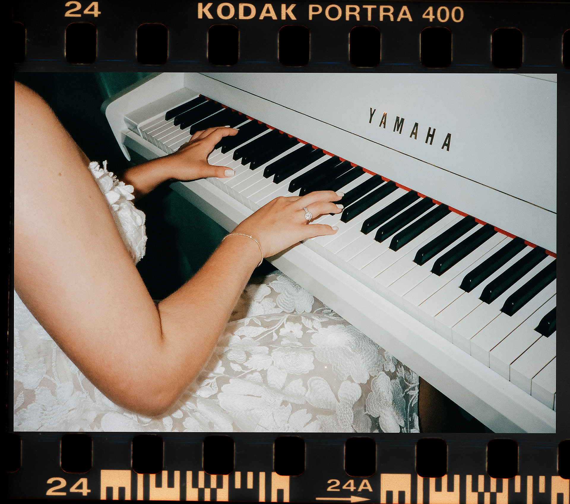 Skan z kliszy Kodak Portra 400 przedstawiający dłonie Panny Młodej w koronkowej sukni ślubnej grającej na białym fortepianie Yamaha, wykonującej niespodziankę dla męża, sfotografowany przez Camera Folks – Wedding Photo & Video.