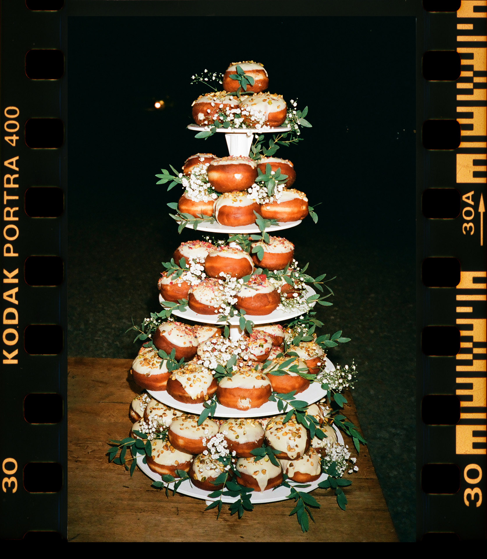 Analogowy skan z kliszy Kodak Portra 400 przedstawiający piętrowy tort weselny w formie piramidy pączków (donutów) z białym lukrem i posypką, ozdobiony zielonymi liśćmi, catering: La Ros Catering (@laroscatering_),